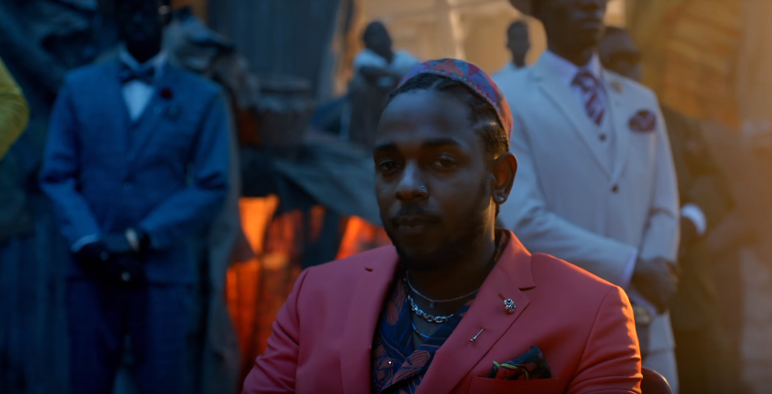 A closeup of Kendrick Lamar sitting in a red tailored suit among brightly-dressed men in a shanty town at sunset.