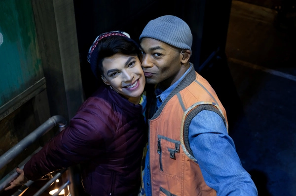 Valentina in boy drag (a maroon puffer coat and maroon an white hat) as Angel and Dixon in a gray ribbed skull cap, orange and grey vest, and blue jean shirt as Tom Collins. Dixon is giving Valentina a peck on the cheek as they pose for the camera. 