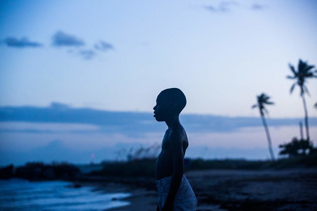 Little Chiron stands on the beach at a blue dusk and looks out at the water. 