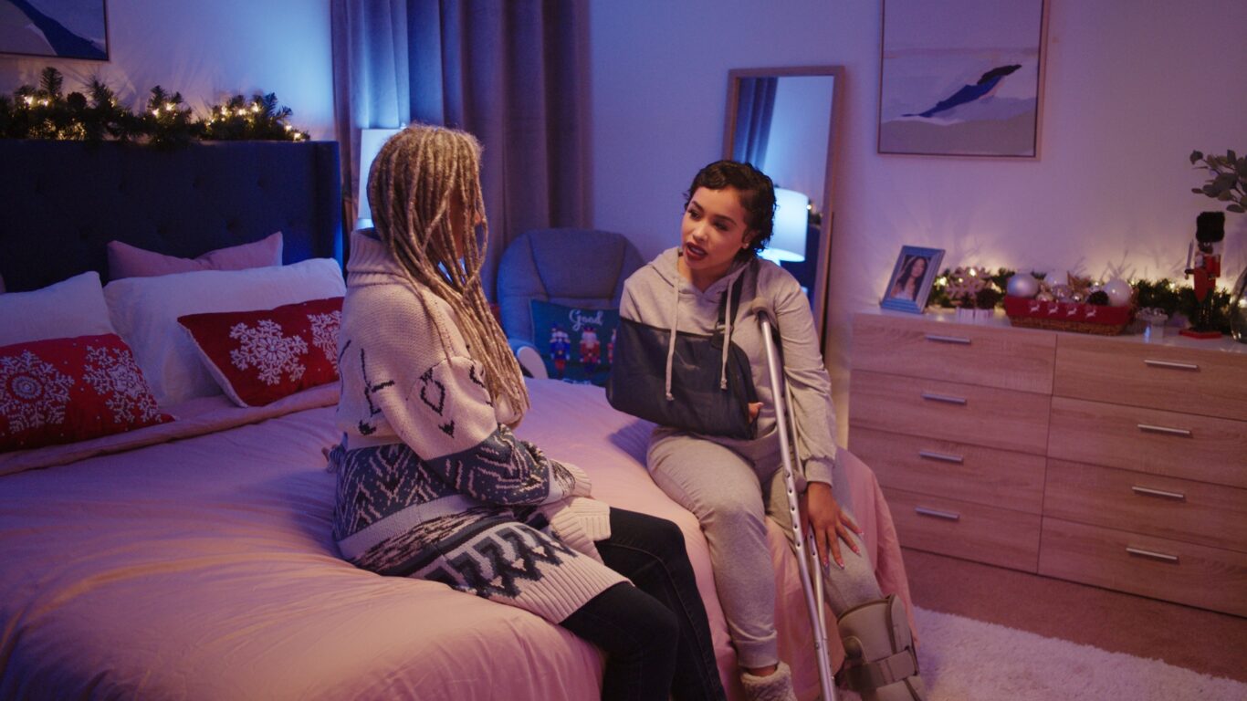 Victoria Rowell and Tabatha Robinson, who is wearing a cast and sling on her arm, are sitting on a festive bed.