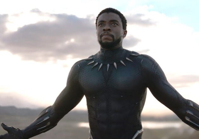 T'Challa, in his Black Panther suit without his mask, walks forward, arms slightly outstretched, in the open landscape of Wakanda.