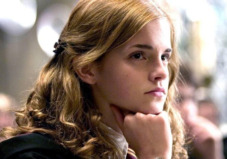 Hermione Granger rests her chin on her closed fits as she looks into the distance while sitting at a table in Hogwarts.