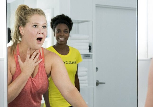 Amy Schumer reacts to her reflection in the gym bathroom mirror with Sacheer Zamata reacting to Schumer in the background in "I Feel Pretty"