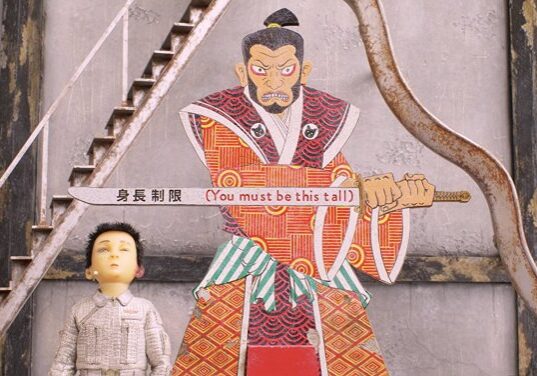 Atari (Koyu Rankin) stands under a Samurai sign that reads "You Must Be This Tall"