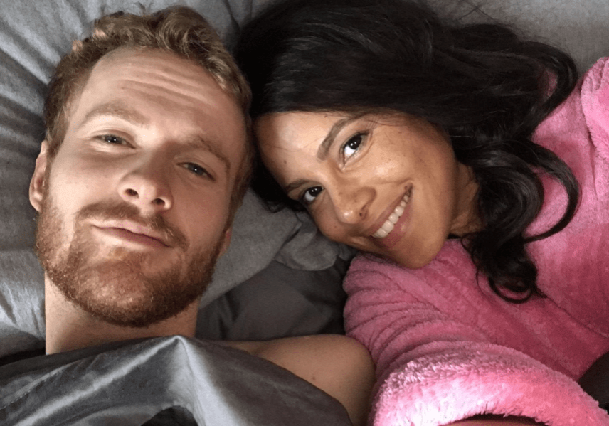 Parisa Fitz-Henley, right, and Murray Fraser take a selfie in costume as Prince Harry and Meghan Markle while lying down on a bed.