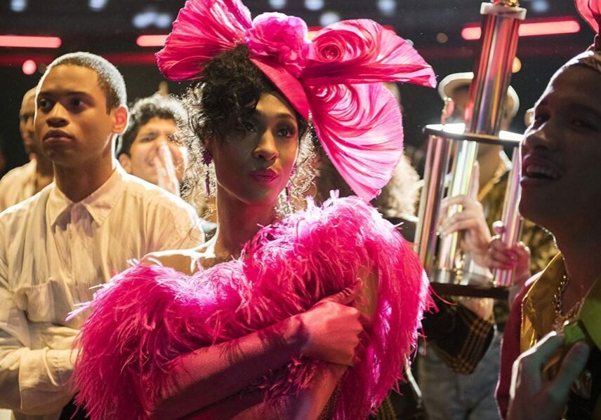 Amid a crowd, Mj Rodriguez, in a feathery pink outfit and wearing a pink hat, holds a trophy after winning a ball category.