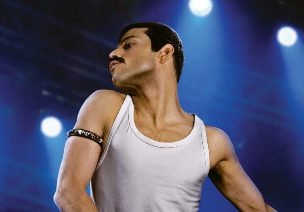 Rami Malek posed as Freddie Mercury in a white tank top and black, spiked armband. He's in front of a blue concert-lit background.