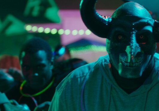 A person in a scary horned mask is in the foreground as black men are possibly partying amid a disturbing color scheme.